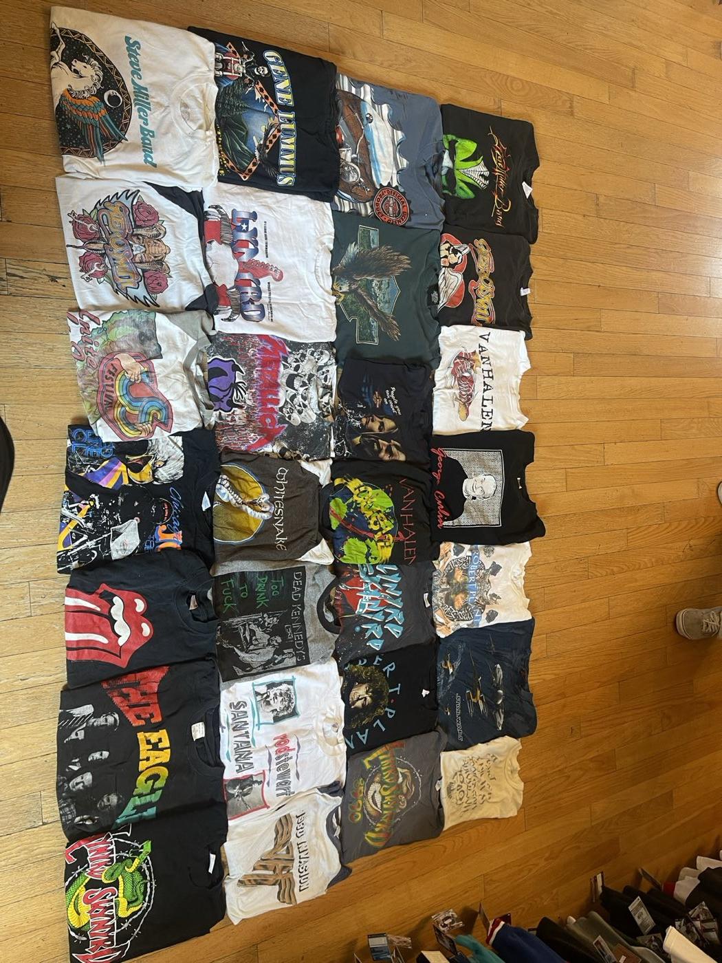 Collection of roughly thirty vintage band t-shirts laid out on a wood floor, including Van Halen, Metallica, and Rolling Stones