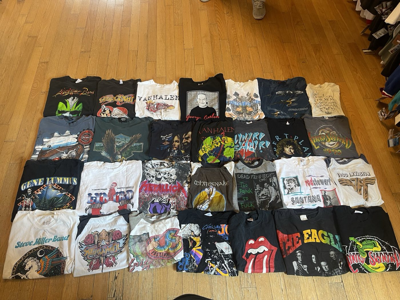 Collection of roughly thirty vintage band t-shirts laid out on a wood floor, including Van Halen, Metallica, and Rolling Stones