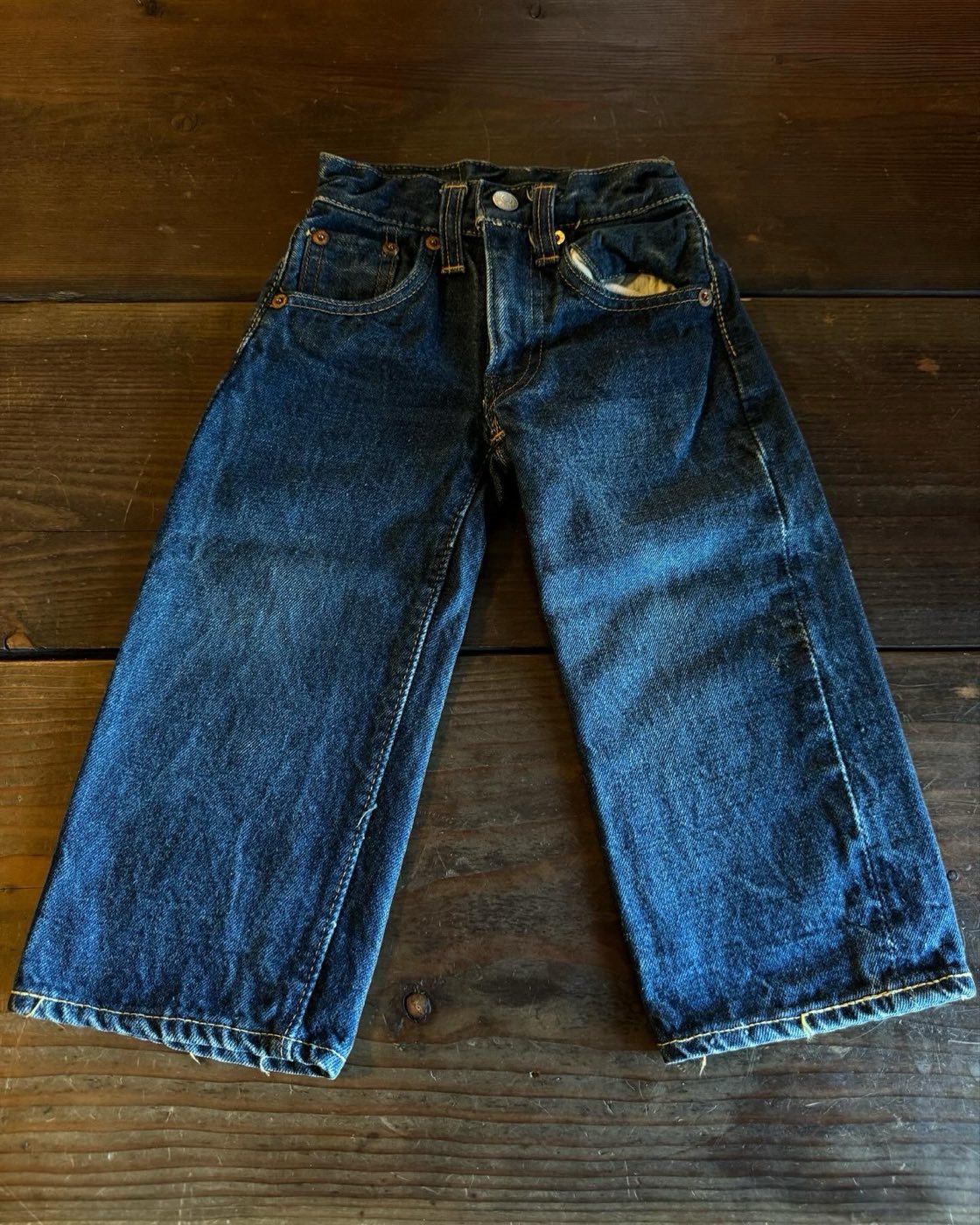 Small dark indigo vintage Levi's jeans laid flat on wooden boards, front view, with copper rivets at the pocket corners, a watch pocket, and light wear at the thighs