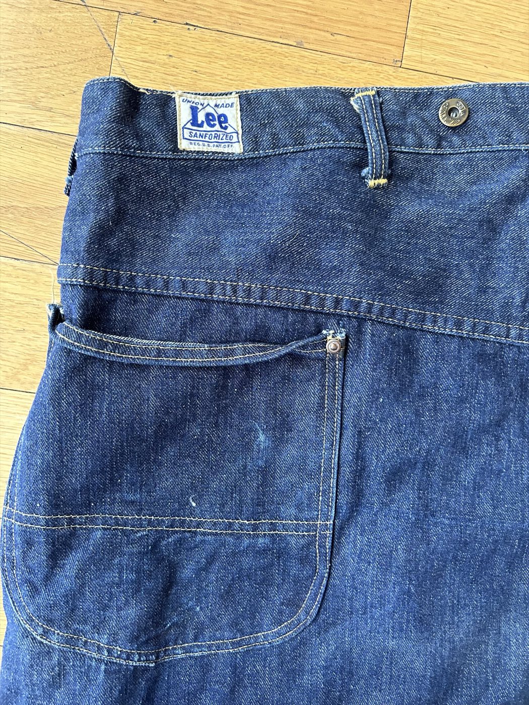 Rear waistband of dark indigo Lee carpenter pants with a white tag reading Union Made, the Lee name inside a house-shaped outline, Sanforized and Reg. U.S. Pat. Off., beside a suspender button and a riveted back pocket