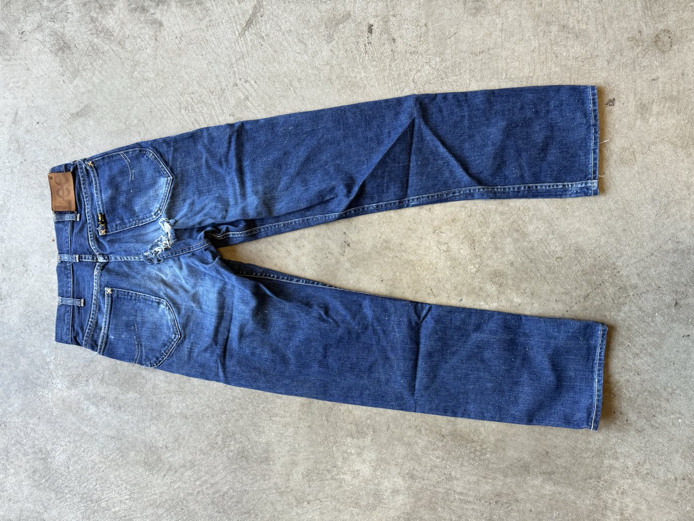 Back of worn vintage Lee Riders jeans laid flat on concrete, showing the leather Lee patch on the waistband, a small black tag on the right back pocket, faded lazy S stitching and a blown-out seat