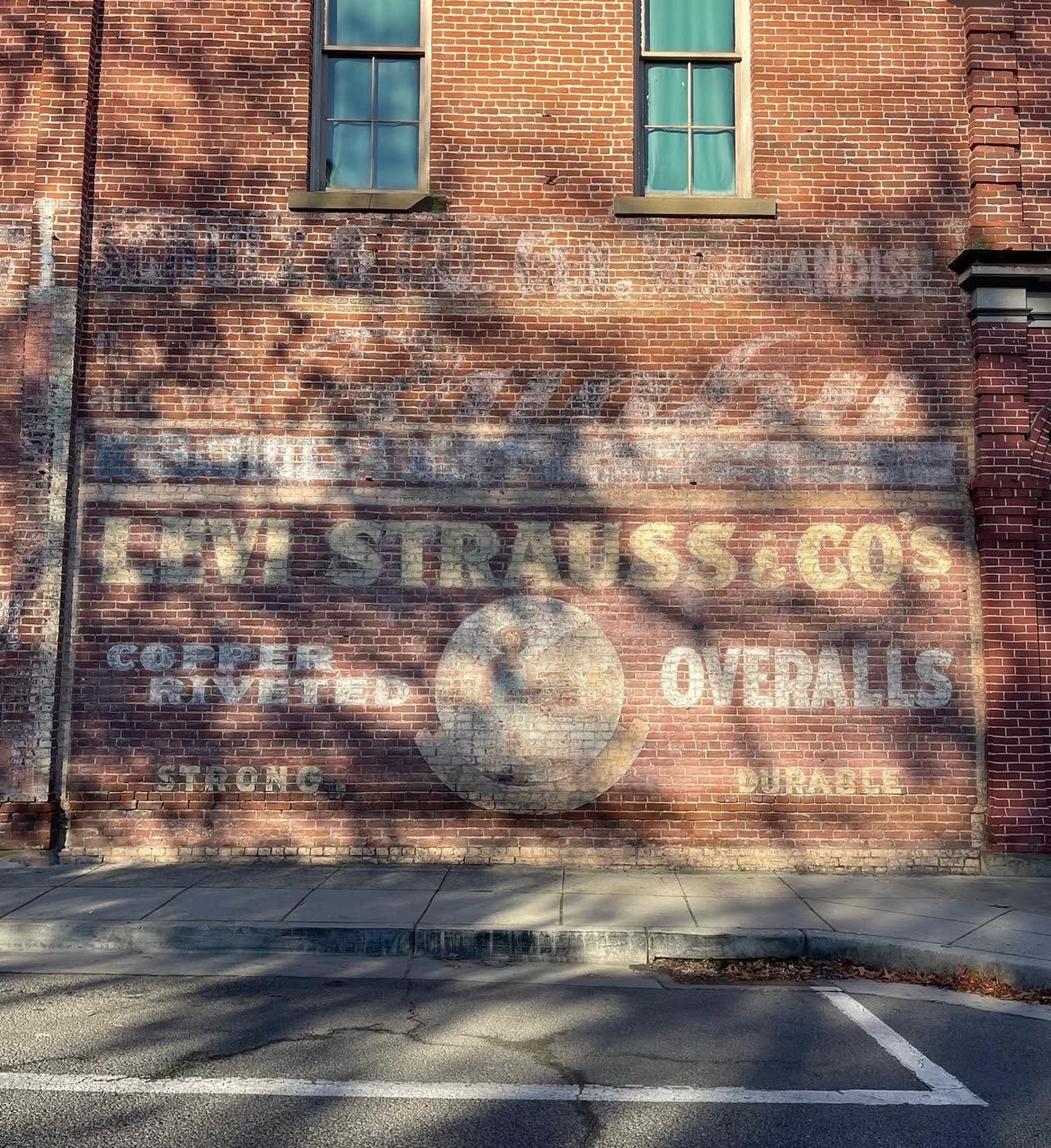 Faded painted Levi Strauss and Co’s Copper Riveted Overalls advertisement on a red brick wall in Jacksonville, Oregon, with a store name and General Merchandise above it, two windows over the sign and a sidewalk below