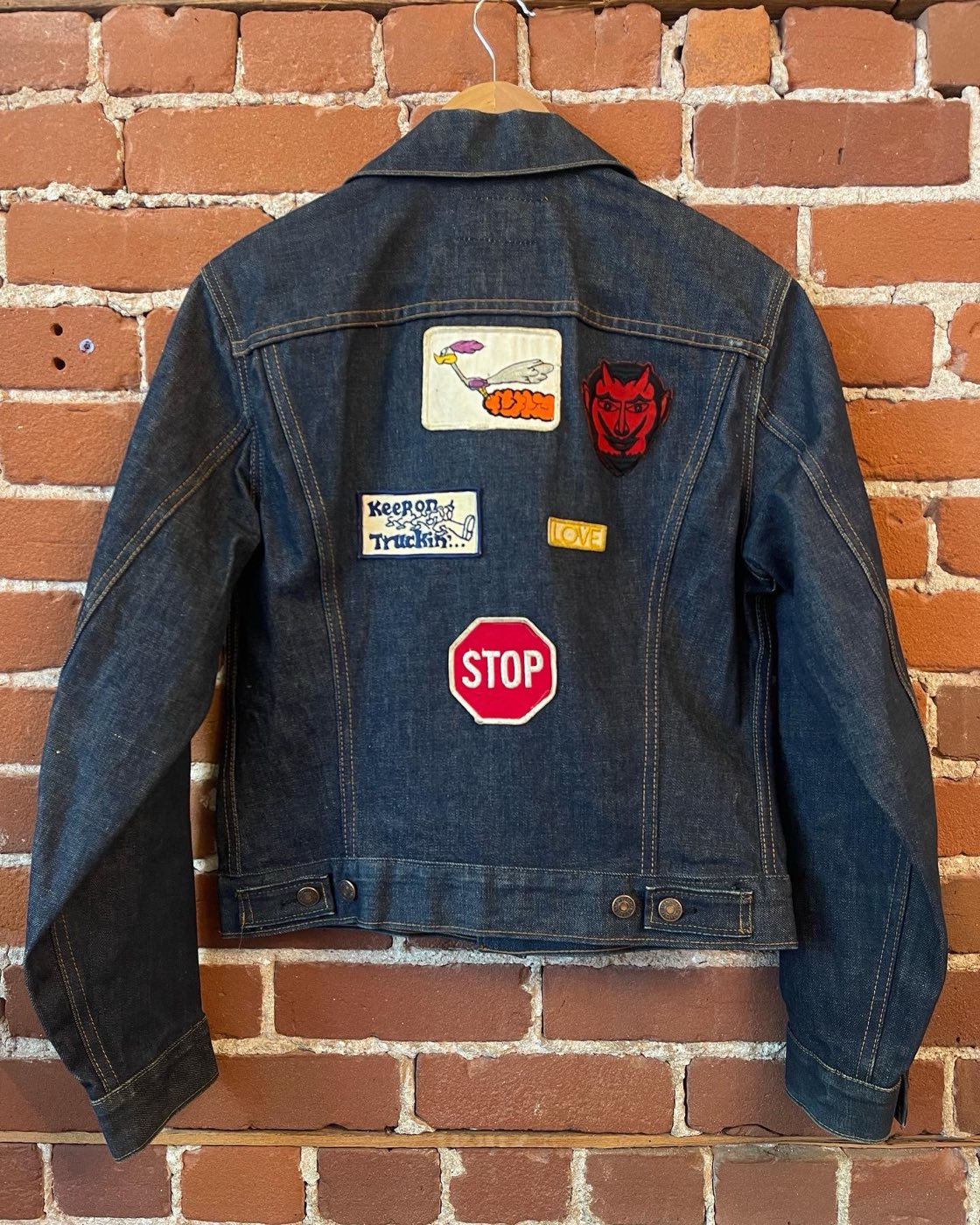 Back of a dark early 1970s Levi's trucker jacket on a hanger covered in period patches including a road runner, a red devil, Keep on Truckin, LOVE, and a stop sign