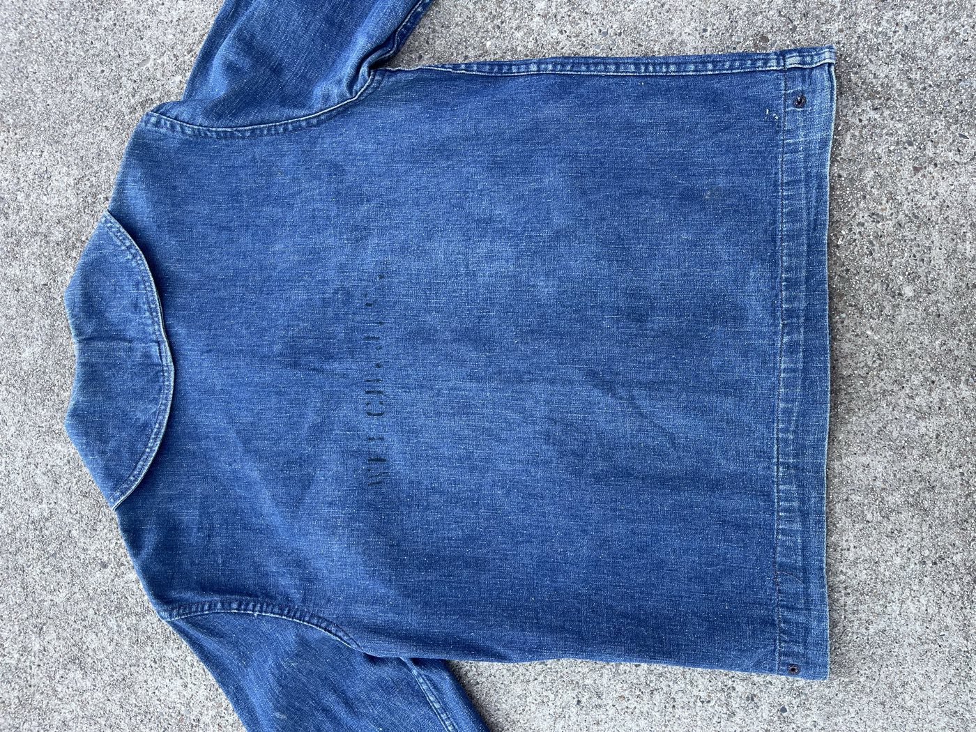 Back of a WWII US Navy denim jacket laid flat, uneven indigo fading across the shoulders, a faint stenciled name down the center, and a metal eyelet near the side hem