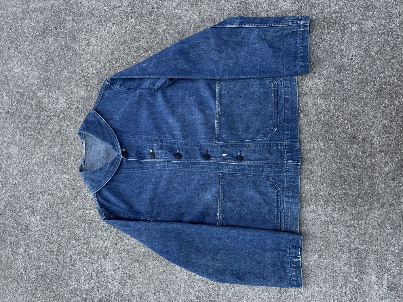 WWII US Navy shawl collar denim jacket laid flat on concrete, five dark buttons down the front, patch pockets at the chest and hip, paint flecks on one cuff